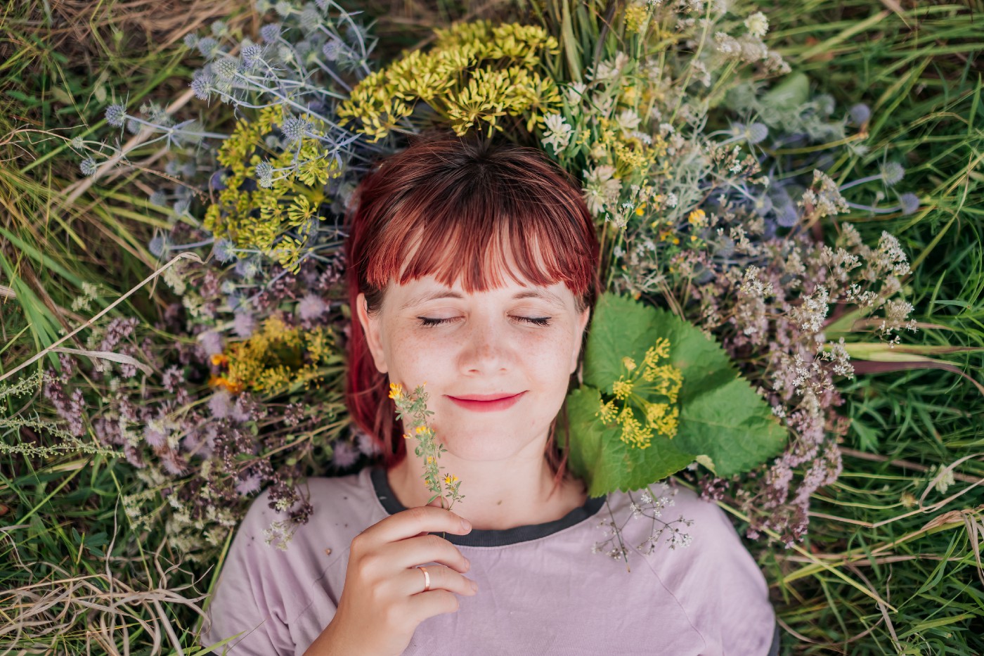 A woman is reclined on the grass with her head surrounded by wild flowers. She is smiling with her eyes closed and holding a sprig of a plant.