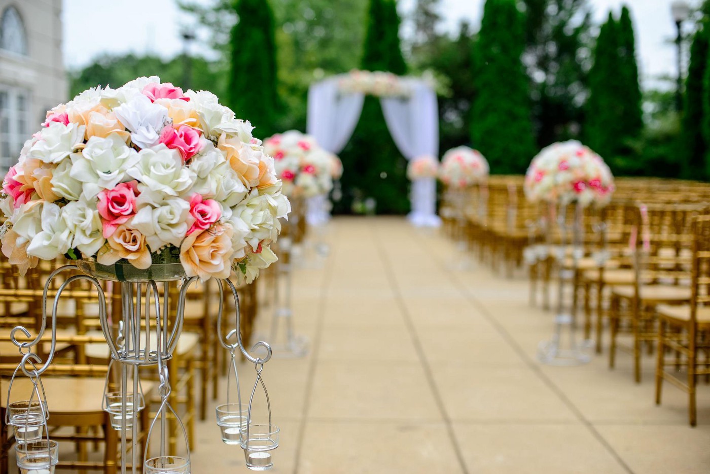 Outdoor wedding setup with center aisle and rows of wooden chairs. A cloth draped arch is at the end of the aisle and large bouquets of flowers are placed on some end chairs.